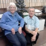 Martha Wachsmuth and Father Bob Antonelli seated in front of a tabletop Christmas tree.