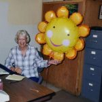Martha Wachsmuth sitting in a chair and smiling while holding a large inflatable sunflower smiley face balloon.