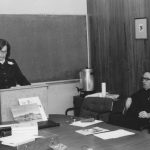 Martha Wachsmuth speaking from a lectern at a table as Father Barry Hagan listens while sitting in a chair nearby.