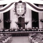 Rows of chairs facing a stage decorated with the University of Portland Crest, ribbons, floral arrangements, and eight chairs across the stage.