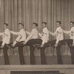 Row of 10 men in letter sweaters with one leg kicked up in an chorus line.