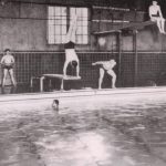 Students diving or getting ready to dive from the edge of an indoor swimming pool.