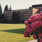 West side view of Buckley Center, large trees to the left and pink rhododendrons in the foreground.