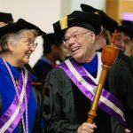 Two faculty Grand Marshals in academic regalia, sash, and each holding a mace.