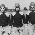 Three football players standing side by side and wearing football helmets, football jersey, and loose pants.