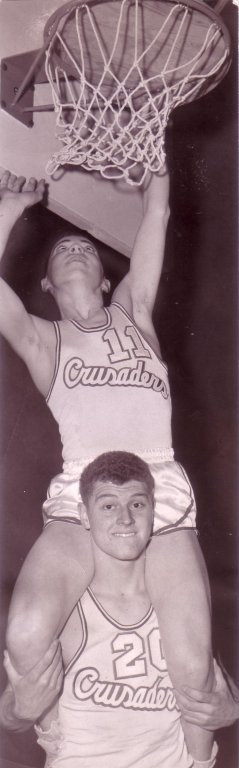 Basketball player carrying another basketball player on his shoulders while standing beneath a basketball hoop.