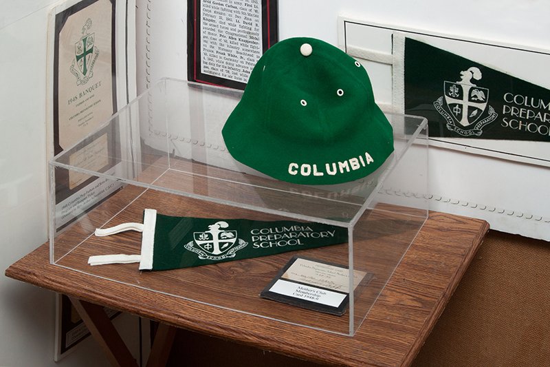 Table display of a green beanie with the word Columbia printed on the brim, Columbia Preparatory School pennant, and a membership card.
