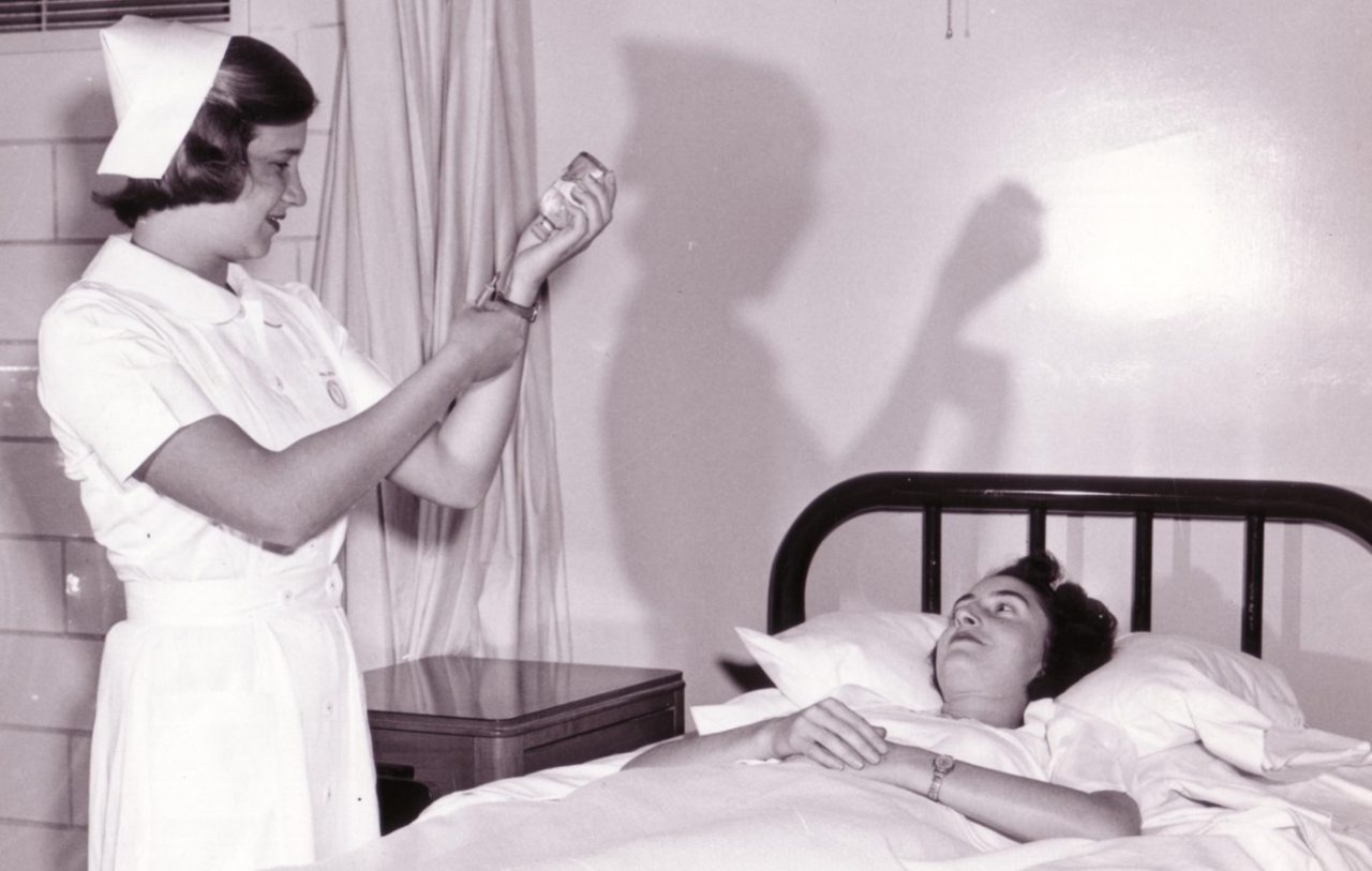 Nurse standing next to a bed with a patient and using a syringe to extract medicine from a bottle.