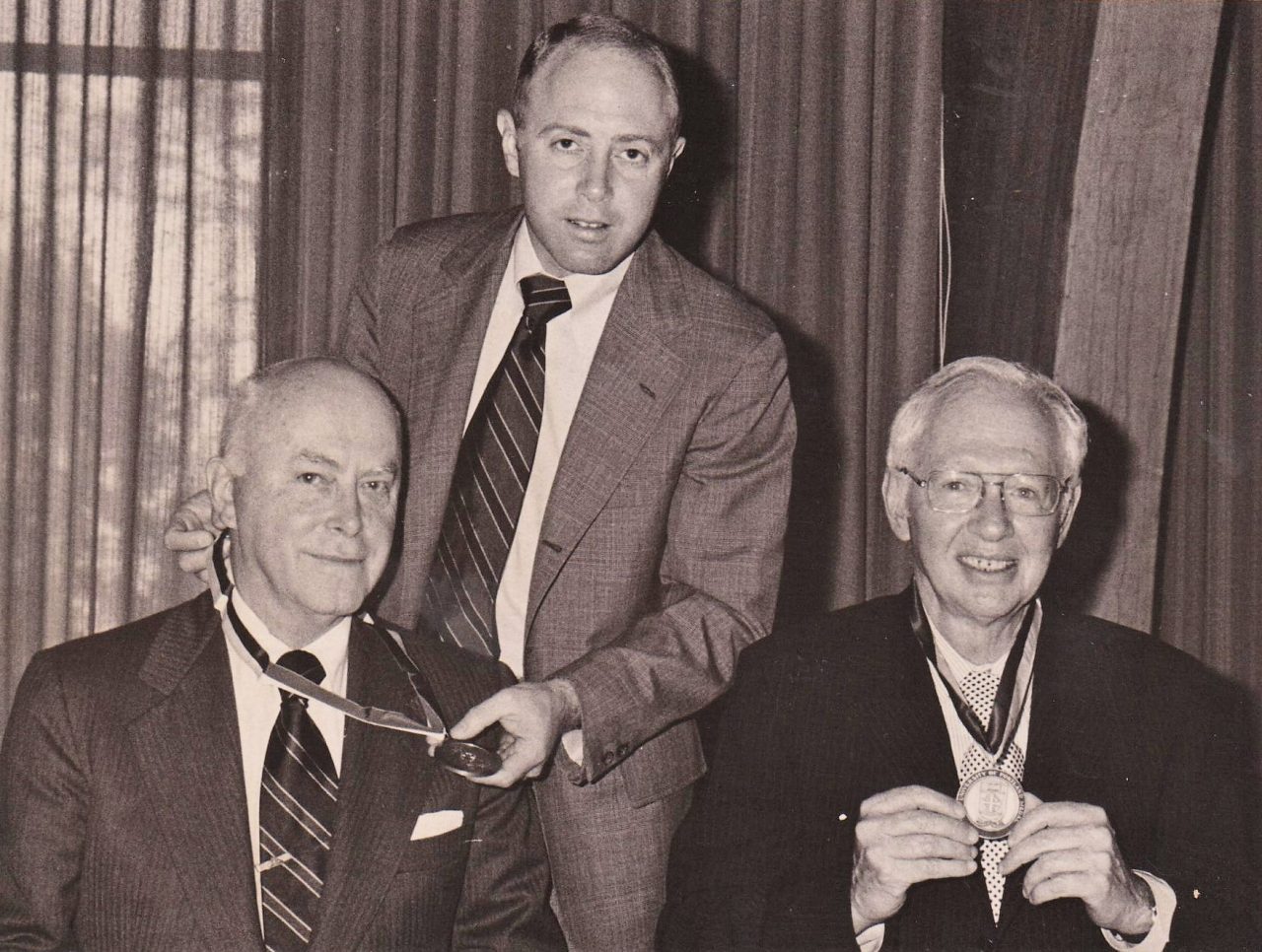 Man standing behind two individuals who are wearing an award medal around their necks.